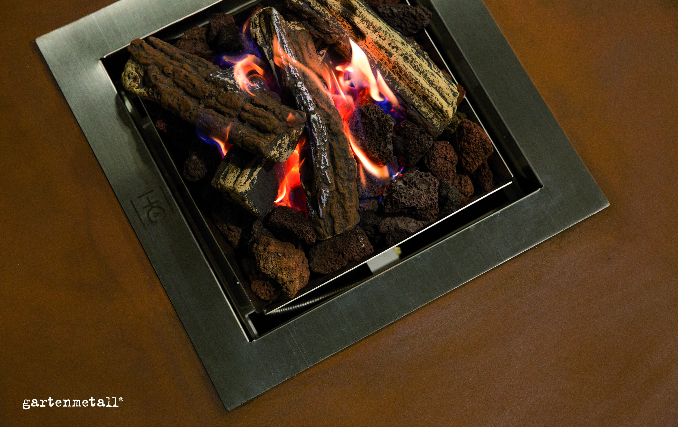 Ein moderner Feuergrill brennt mit rötlichen Flammen auf künstlichen Holzscheiten und Lavasteinen, umrahmt von einem Edelstahlrahmen auf einer braunen Oberfläche. Text: 'gartenmetall®'.
