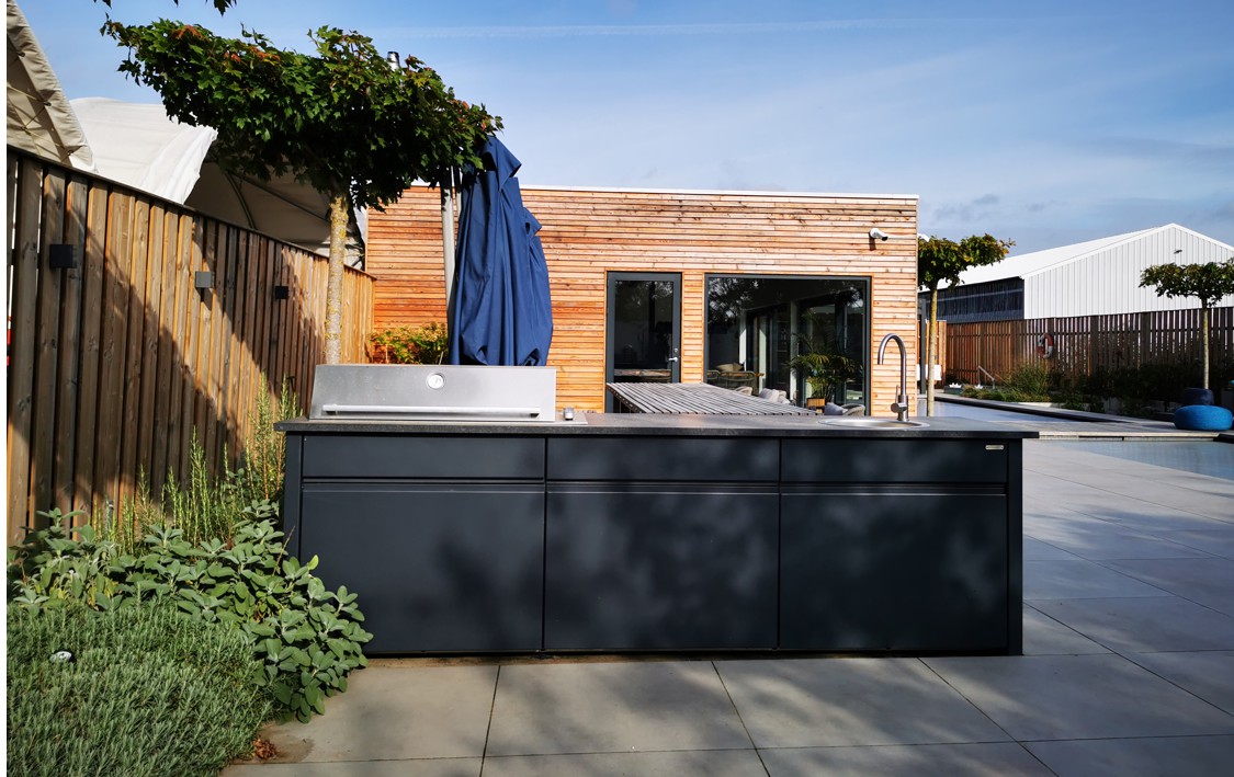 Eine dunkle Outdoor-Küche steht auf einer Terrasse neben einem Sonnenschirm. Im Hintergrund ist ein modernes Holzgebäude mit Glasfronten. Pflanzen säumen den Bereich entlang eines Holzzauns.