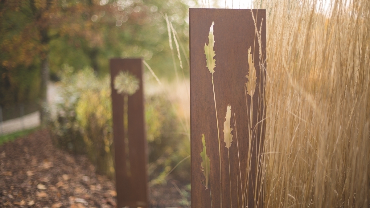 Metallplatte steht, zeigt eingearbeitete Pflanzenmotive, umgeben von hohen, goldenen Gräsern im unscharfen herbstlichen Garten.