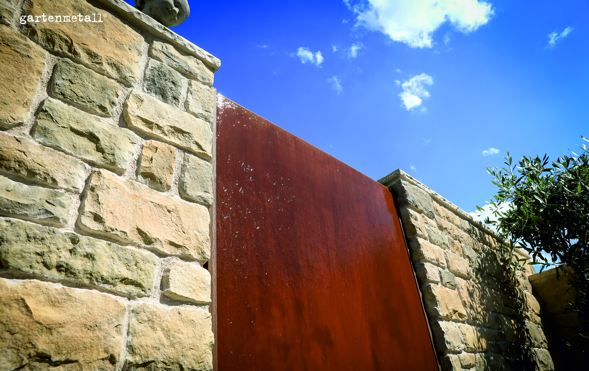 Steinmauer steht neben rostfarbener Metallplatte, umgeben von blühendem Baum unter klarem, blauem Himmel. Text: 'gartenmetall' oben links.