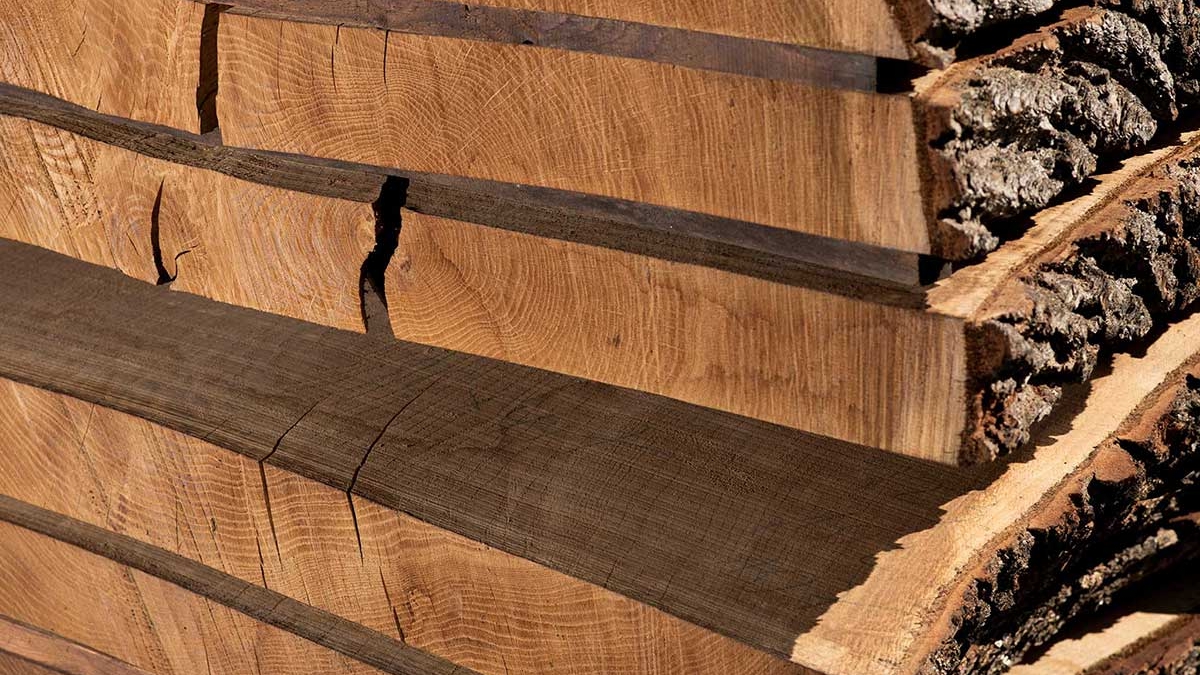 Gestapelte Holzbohlen ruhen in der Sonne, ihre Rinde teils sichtbar, in einem Außenbereich. Die Maserungen und Trockenrisse zeugen von Natur und Zeit.