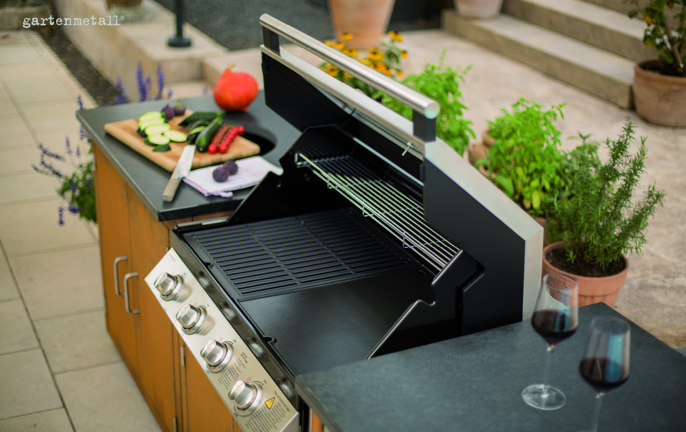 Grill mit geöffnetem Deckel steht unbenutzt auf einer Terrasse, umgeben von Pflanzen. Schneidebrett mit Gemüse und Messer daneben. Zwei gefüllte Weingläser stehen auf dem Tisch. Text: 'gartenmetall'.