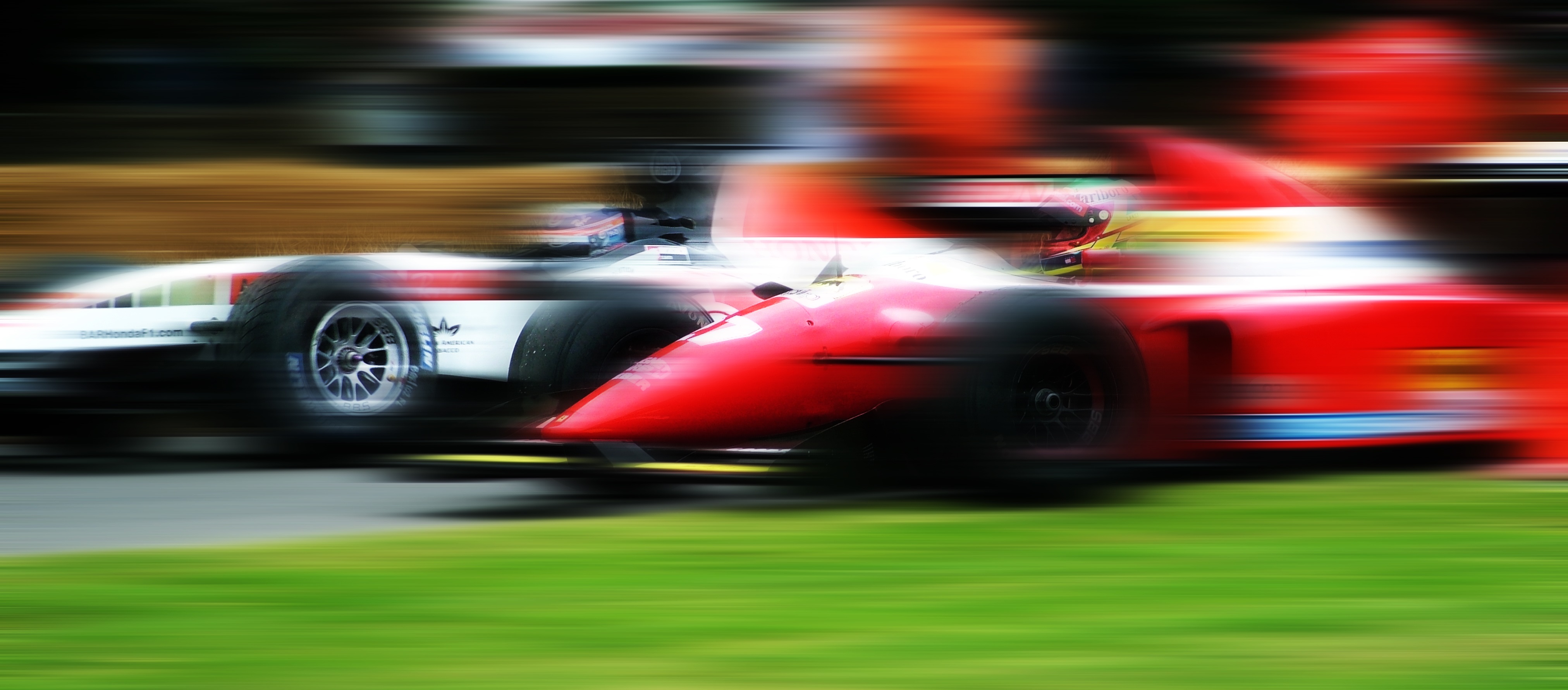 Rennwagen fahren schnell auf einer Rennstrecke. Die Fahrzeuge erscheinen verwischt durch die Geschwindigkeit. Im Hintergrund sind verschwommene Zuschauer zu sehen. Aufschrift: 'BARHondaF1.com'. Gras grenzt die Strecke ab.