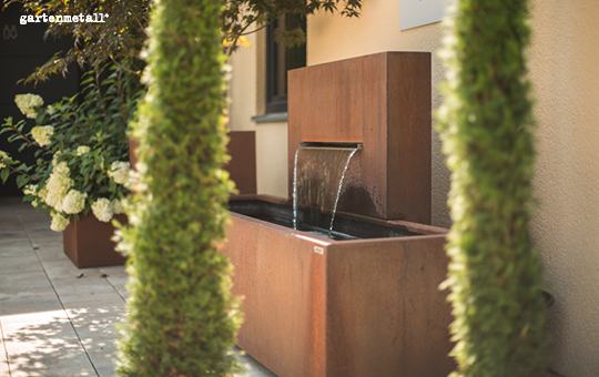 Ein rechteckiger Metallbrunnen fließt in einem modernen Garten, umgeben von grünen Pflanzen und weißen Blüten. Die Schrift im Bild lautet: 'gartenmetall.'