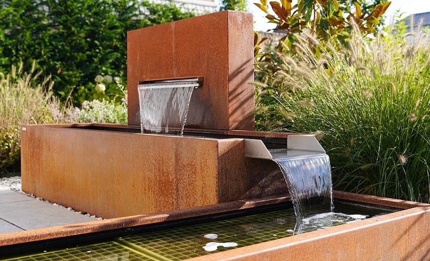 Ein rechteckiger Springbrunnen aus rostigem Metallfließt Wasser kaskadenartig, umgeben von grünen Pflanzen und Gräsern in einem Garten bei Tageslicht.