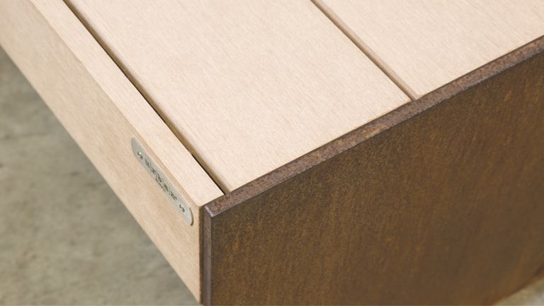 Holzbox, ruht auf einem Betonboden, zeigt eine metallene Namensplakette mit der Aufschrift 'CREABETON'. Die Box hat eine rostige, metallene Kante.