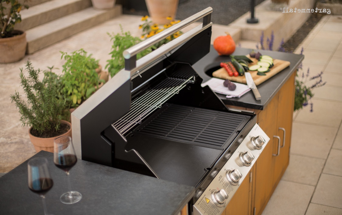 Gasgrill steht geöffnet auf Terrasse, auf seitlicher Ablage Holzbrett mit geschnittenem Gemüse, Messer, daneben zwei Rotweingläser auf schwarzer Tischplatte, umgeben von Pflanzen in Töpfen.