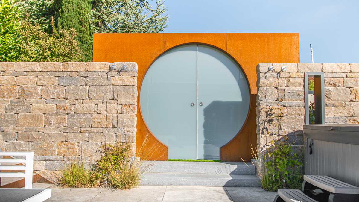Runde Glastür steht geschlossen in einer rostfarbenen Mauer, flankiert von Steinwänden. Davor befinden sich Stufen, Büsche und Gräser, alles in einem sonnigen Garten.
