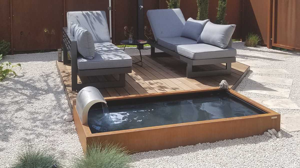 Gartenlounge mit grauen Polstern auf Holzterrasse, neben rechteckigem Wasserbecken mit dekorativem Wasserfall, umgeben von Kieselsteinen und Holzzaun, sonnige Atmosphäre.