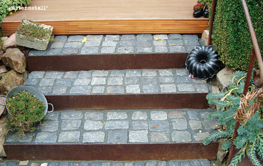 Steintreppen führen hoch, flankiert von Pflanzen in Töpfen und Ziegeln. Ein dunkelglänzendes Formobjekt liegt auf den Stufen. Kontext bildet eine hölzerne Plattform. Text: 'gartenmetall®'.