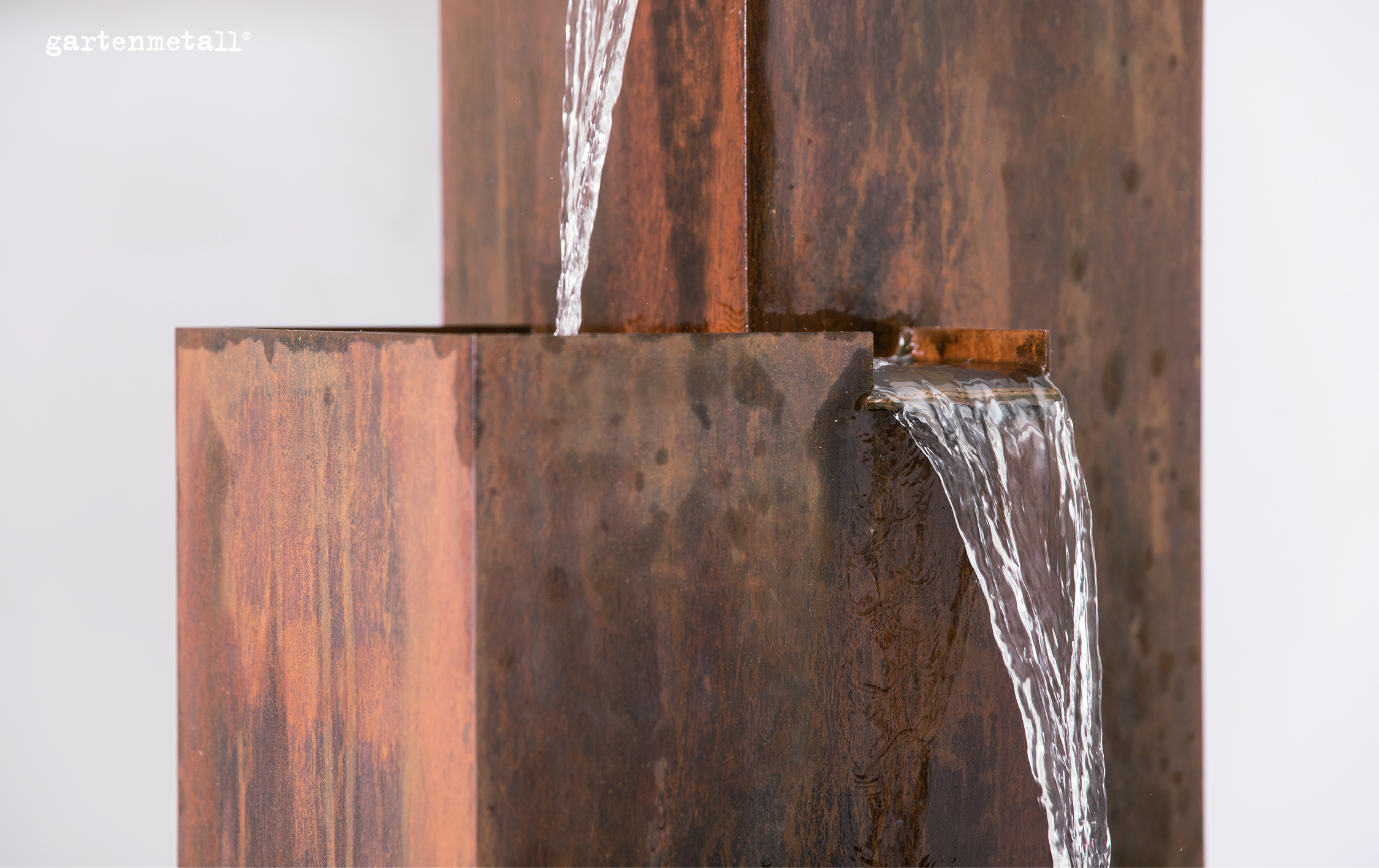 Rostige Metallskulptur lässt Wasser in zwei Strömen fließen, umgeben von minimalistischer, heller Umgebung, oben links Text: 'gartenmetall®'.