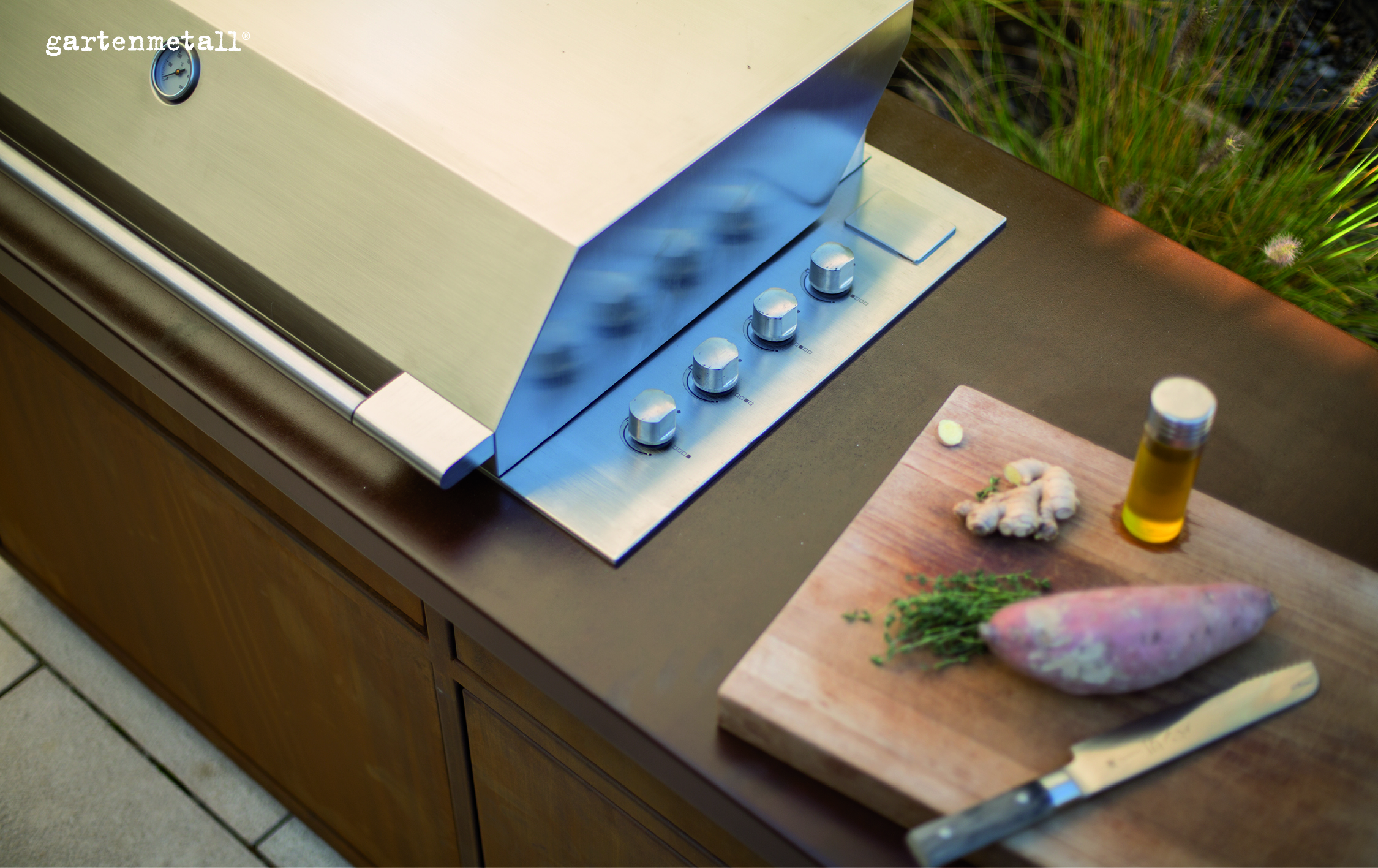 Ein Edelstahlgrill mit Deckel und Knöpfen steht neben einem Schneidebrett mit Ingwer, Süßkartoffel, Thymian und Öl. Im Hintergrund sind Gräser sichtbar. Text: 'gartenmetall®'.