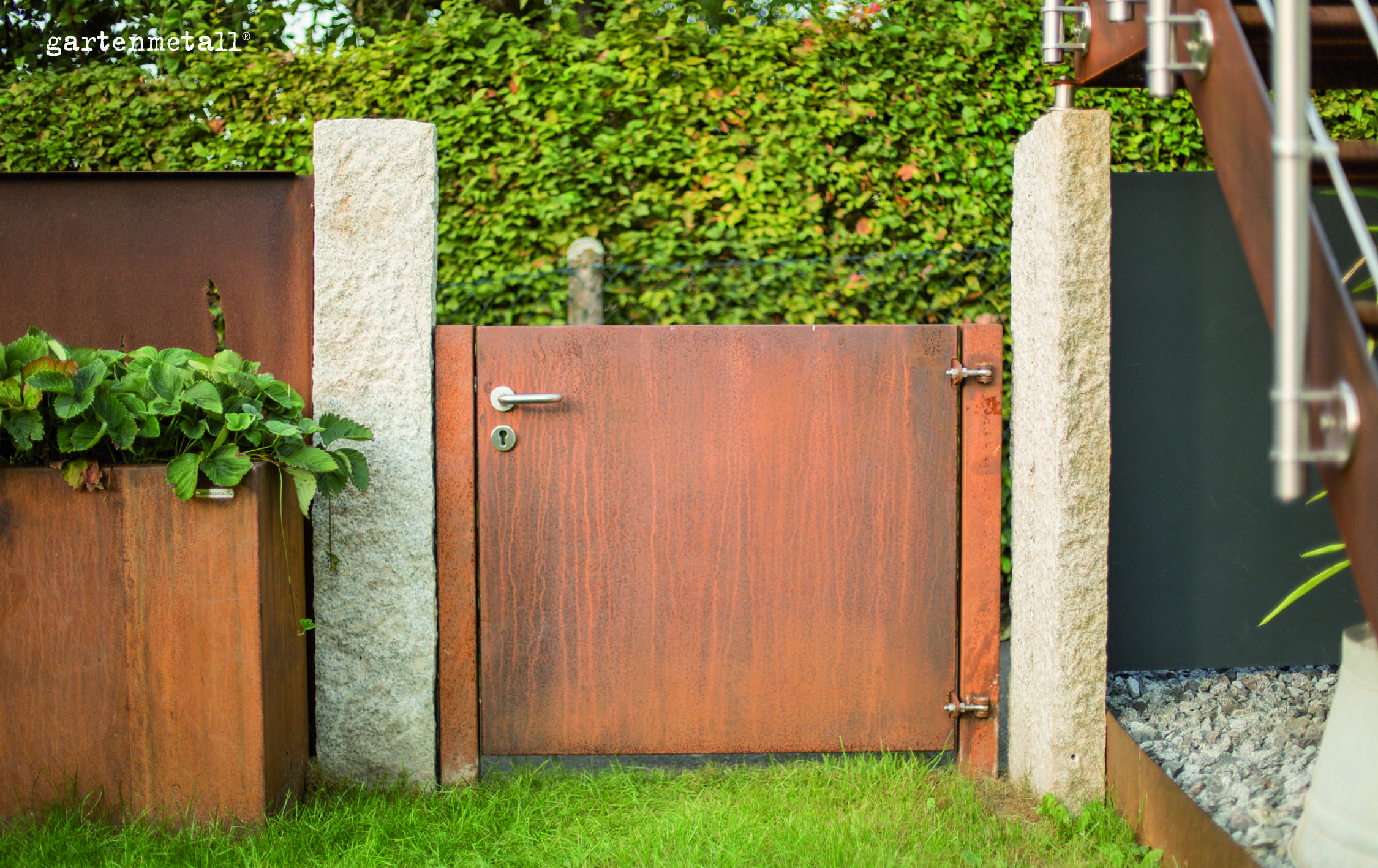Gate of rusty metal, standing closed between two stone pillars. Next to it, plant containers with strawberries, background of hedge and pebbles. Text: 'gartenmetall®'.