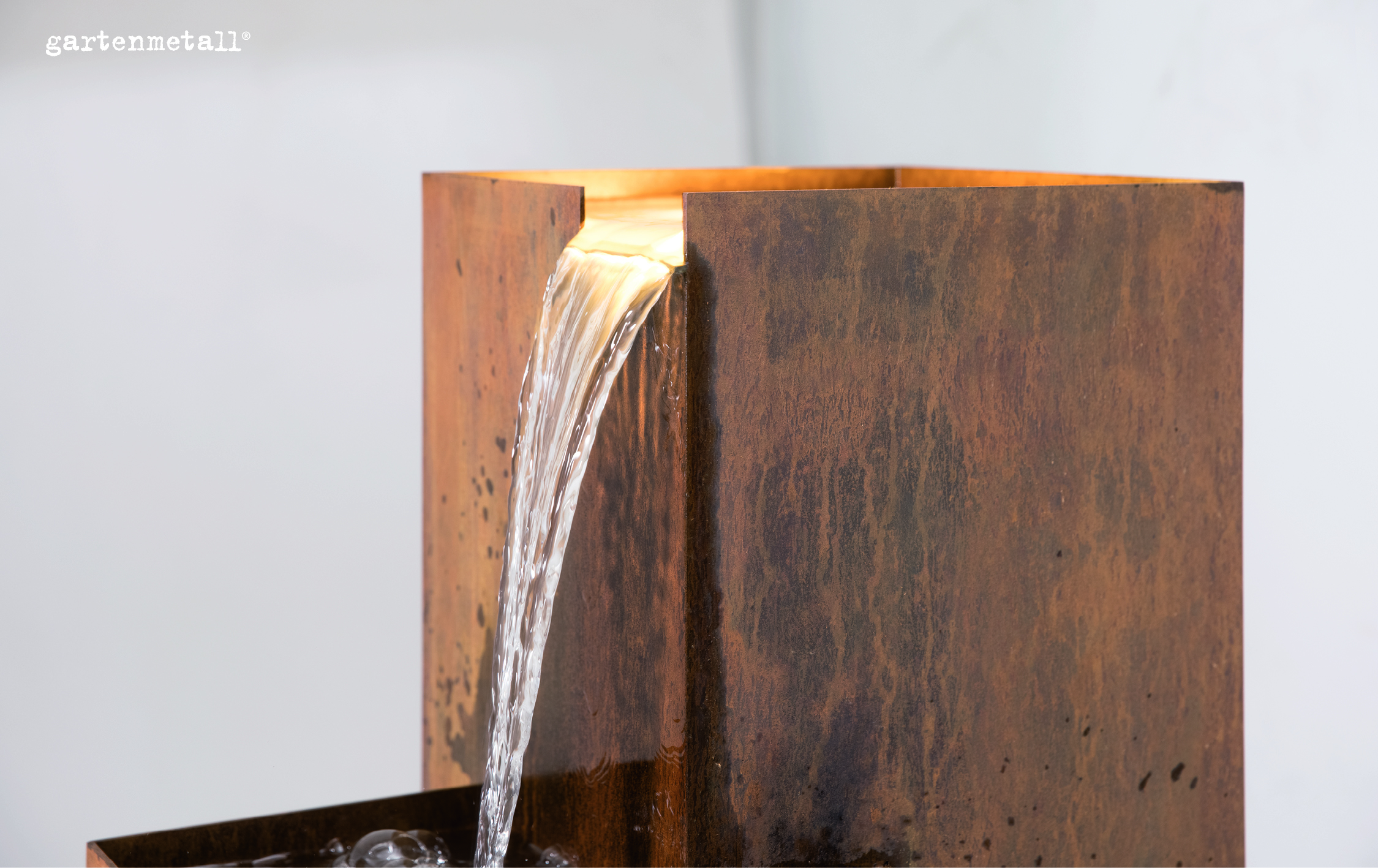 Ein rostfarbener, vertikaler Wasserfall fließt aus einem rechteckigen Metallbehälter in einen darunterliegenden Behälter. Rechts oben steht „gartenmetall®“. Der Hintergrund ist neutral.