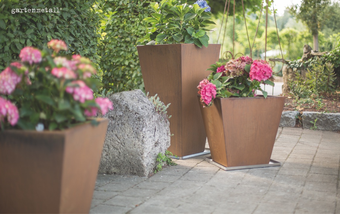 Blumenkübel aus Metall halten bunte Hortensien, die auf einer Terrasse neben einem großen Stein stehen. Im Hintergrund ist ein grüner Garten und der Schriftzug 'gartenmetall®' zu sehen.
