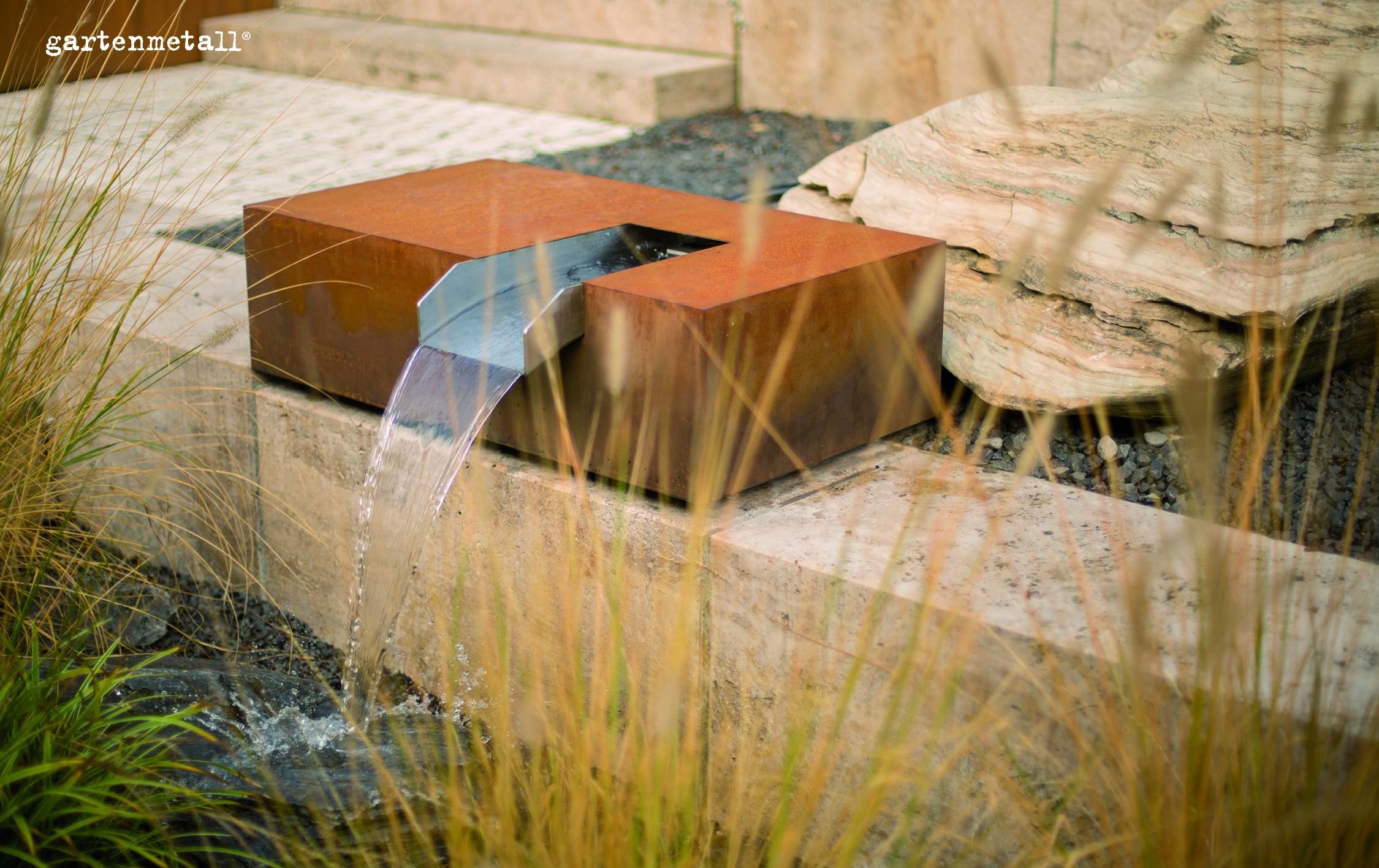 Metallischer Brunnen fließt in klarer Wasserform, umgeben von Steinen und Gras in einem modernen Garten. Text: „gartenmetall®“.