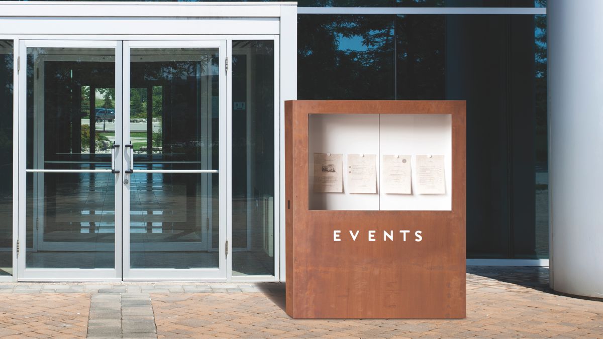 Ein braunes Holzbrett mit der Aufschrift 'EVENTS' zeigt Papierblätter hinter Glas. Steht vor einem modernen Glasgebäude mit doppelten Glas-Schiebetüren.