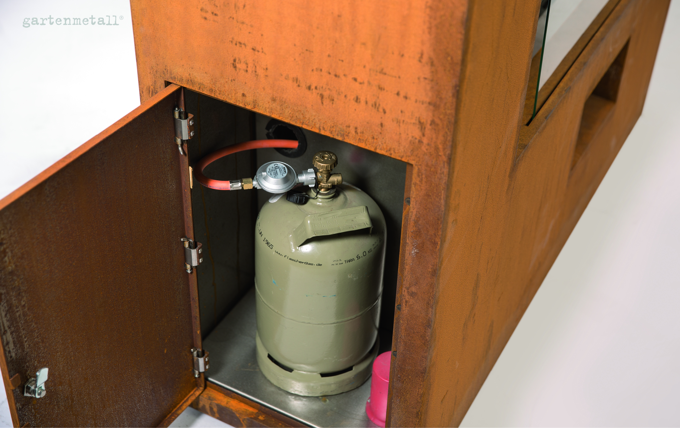Gasflasche steht in einem geöffneten, rostfarbenen Metallschrank, angeschlossen durch einen roten Schlauch. Im Inneren Text: „www.gasheizung.de“. Umgebung ist minimalistisch gestaltet. Text: „gartenmetall®“.