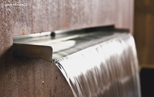 Ein rechteckiger Metallwasserfall fließt gleichmäßig aus einer Wand, umgeben von rostfarbenen Oberflächen. Text vorhanden: 'gasteinmetall'.