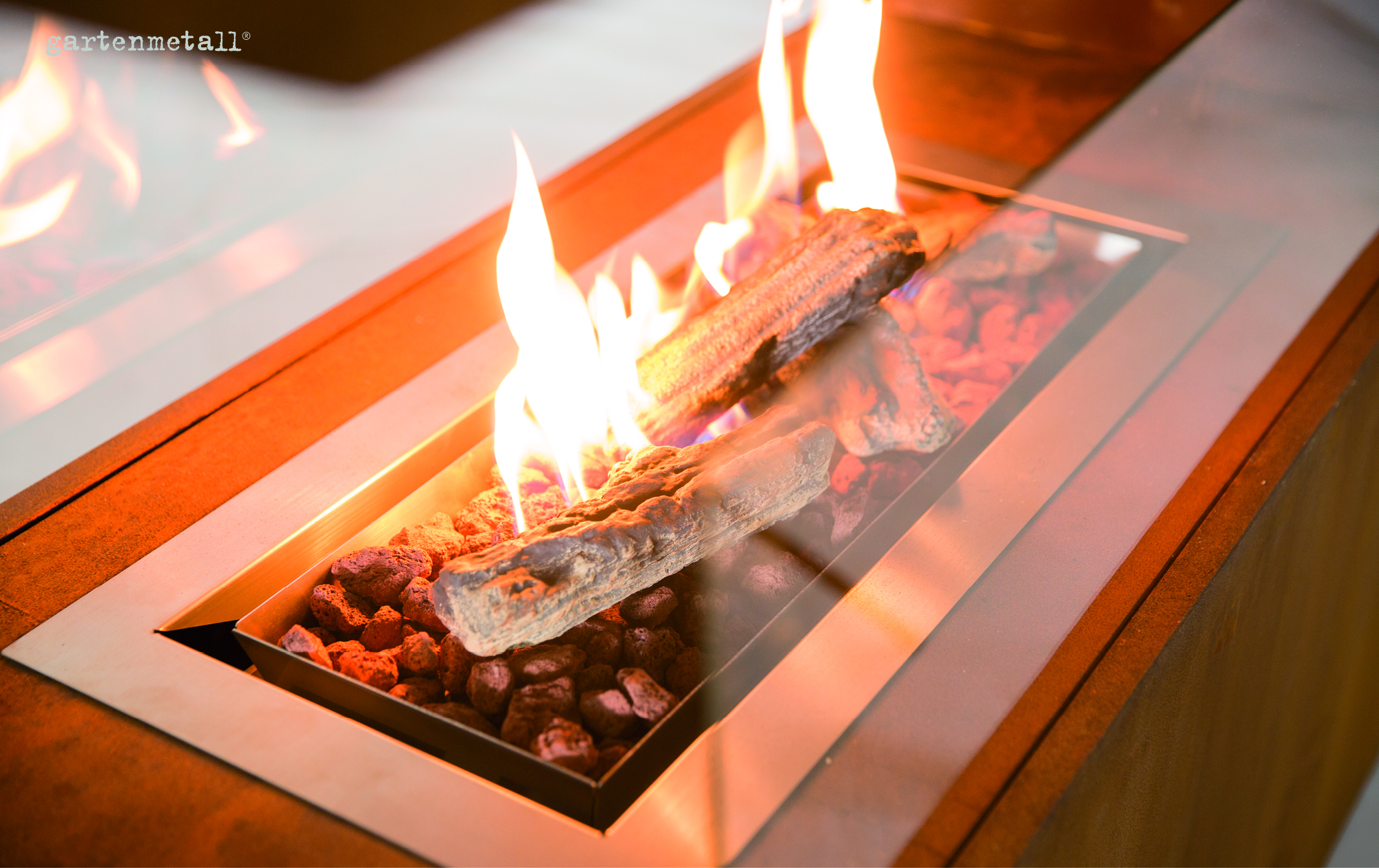 Ein rechteckiger Feuerstelleinsatz brennt mit Flammen über künstlichen Holzscheiten und Lavasteinen, eingeschlossen in einer Metallumrandung. Text: 'gartenmetall®'.