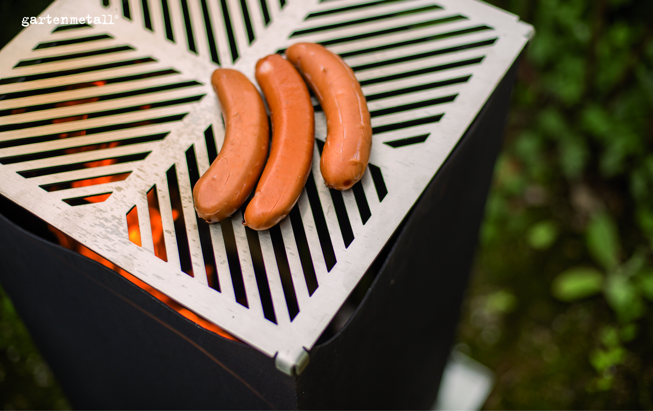 Drei Würste liegen auf einem Grillrost, der über hellen Flammen angebracht ist, im Freien. Text: 'gartenmetall®'.