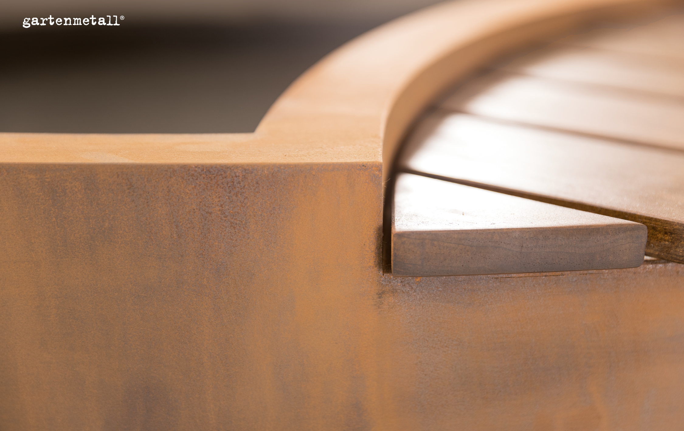 Rostfarbener Metallelement sitzt in geschwungener Form auf Holzuntergrund, nahe Umgebung unscharf. Text: „gartenmetall®“ oben links.