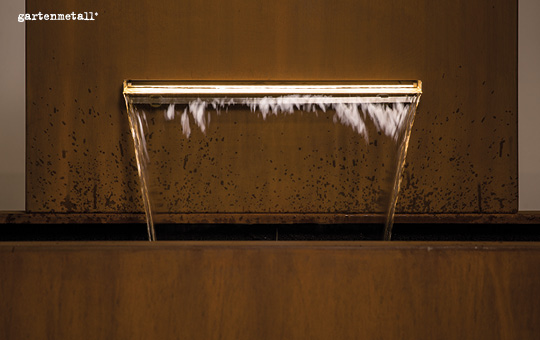 Ein Wasserauslaufbrunnen aus Metall speit Wasser gleichmäßig in einen Auffangbehälter. Die braungefärbte, strukturierte Metalloberfläche trägt die Aufschrift 'gartenmetall®'.