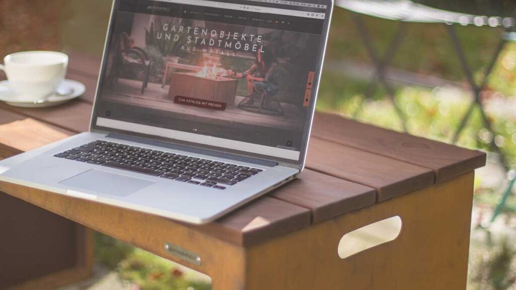 Laptop displaying website 'Garden Objects and Street Furniture.' Placed on a wooden table next to a cup in the garden. Text: 'To the Catalogue with Prices.'