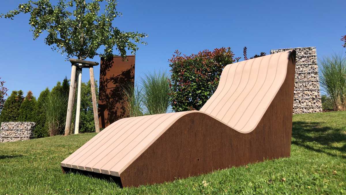 Eine Wellenform-Liege steht ruhig auf einem gepflegten Rasen, umgeben von verschiedenen Pflanzen und einem Baum, unter einem klaren blauen Himmel.