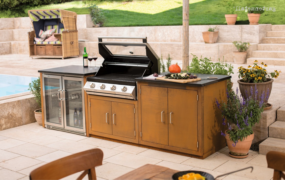 Grillstation mit offenem Deckel steht auf einer Terrasse, flankiert von Kühlschrank und Pflanzen. Schneidebrett mit Gemüse darauf. Zwei Weingläser und Flasche daneben. Text: '1fließentag'.