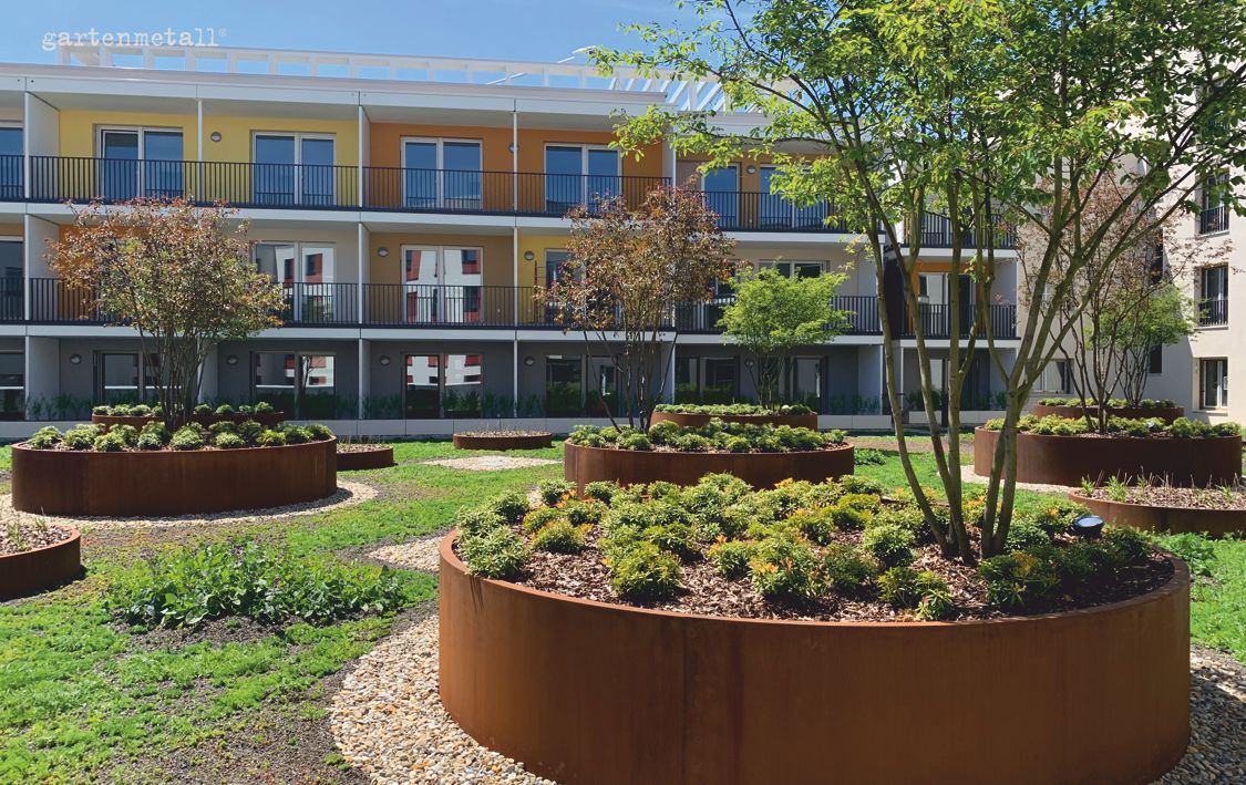 Runde Hochbeete mit Pflanzen stehen auf einer grünen Rasenfläche. Im Hintergrund ist ein modernes, mehrstöckiges Wohngebäude mit bunten Fassaden. Text: 'gartenmetall©'.