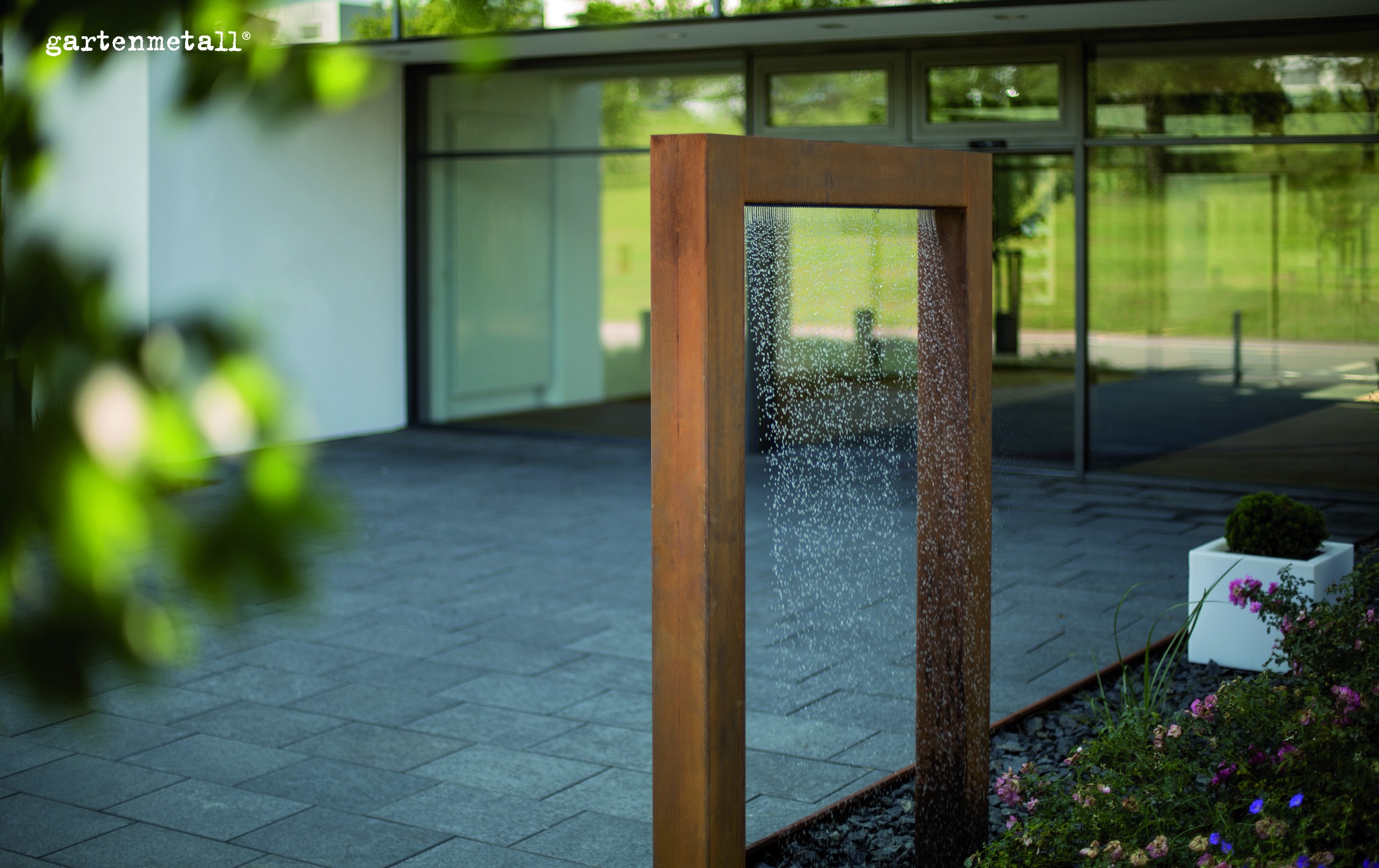 Metallrahmen, durch den Wasser fließt, steht ruhig im gepflasterten Außenbereich neben einem modernen Glasgebäude, grüne Blattpflanzen und Blumen schmücken den Vordergrund. Text: 'gartenmetall'.