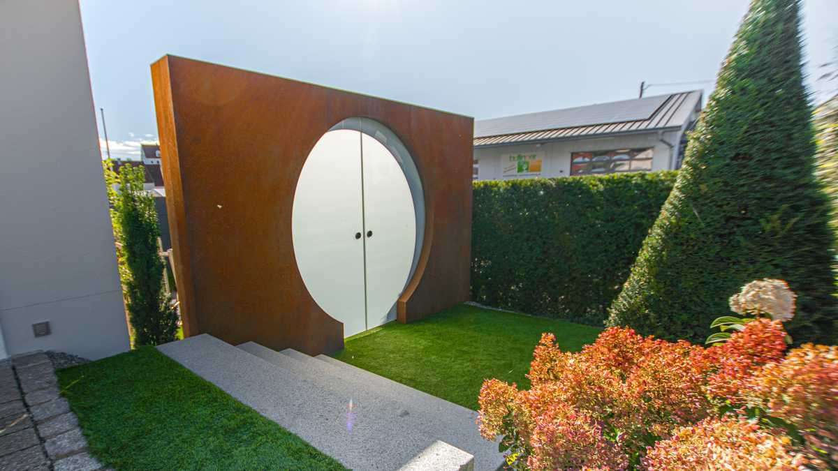 Eine runde, weiße Tür ist in einem rostfarbenen Metallrahmen eingefasst, steht mitten im grünen Garten, umgeben von Hecken und einem modernen Gebäude im Hintergrund.