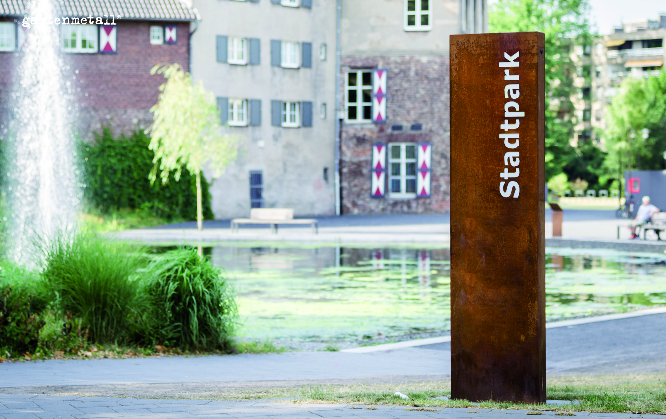 Stele aus rostigem Metall mit der Aufschrift „Stadtpark“ steht vor einem Teich. Im Hintergrund Gebäude und Parkvegetation. Links springender Wasserstrahl, Menschen sitzen im Parkbereich.