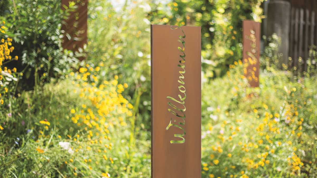 Metallplatte mit eingraviertem „Willkommen“ steht aufrecht in blühendem Garten mit gelben Blumen, umgeben von grünem Laub im Sonnenlicht.
