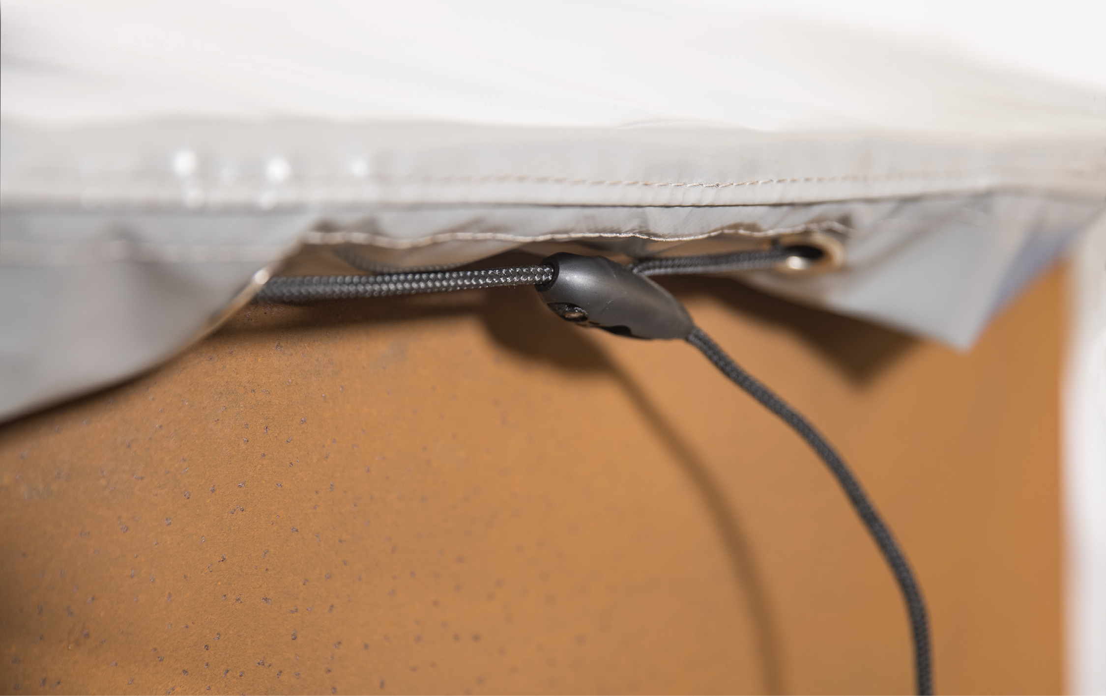 Ein schwarzer Gummizug mit Stopper verläuft durch eine Öse an einer grauen Plane, die über einer rostfarbenen Oberfläche gespannt ist.