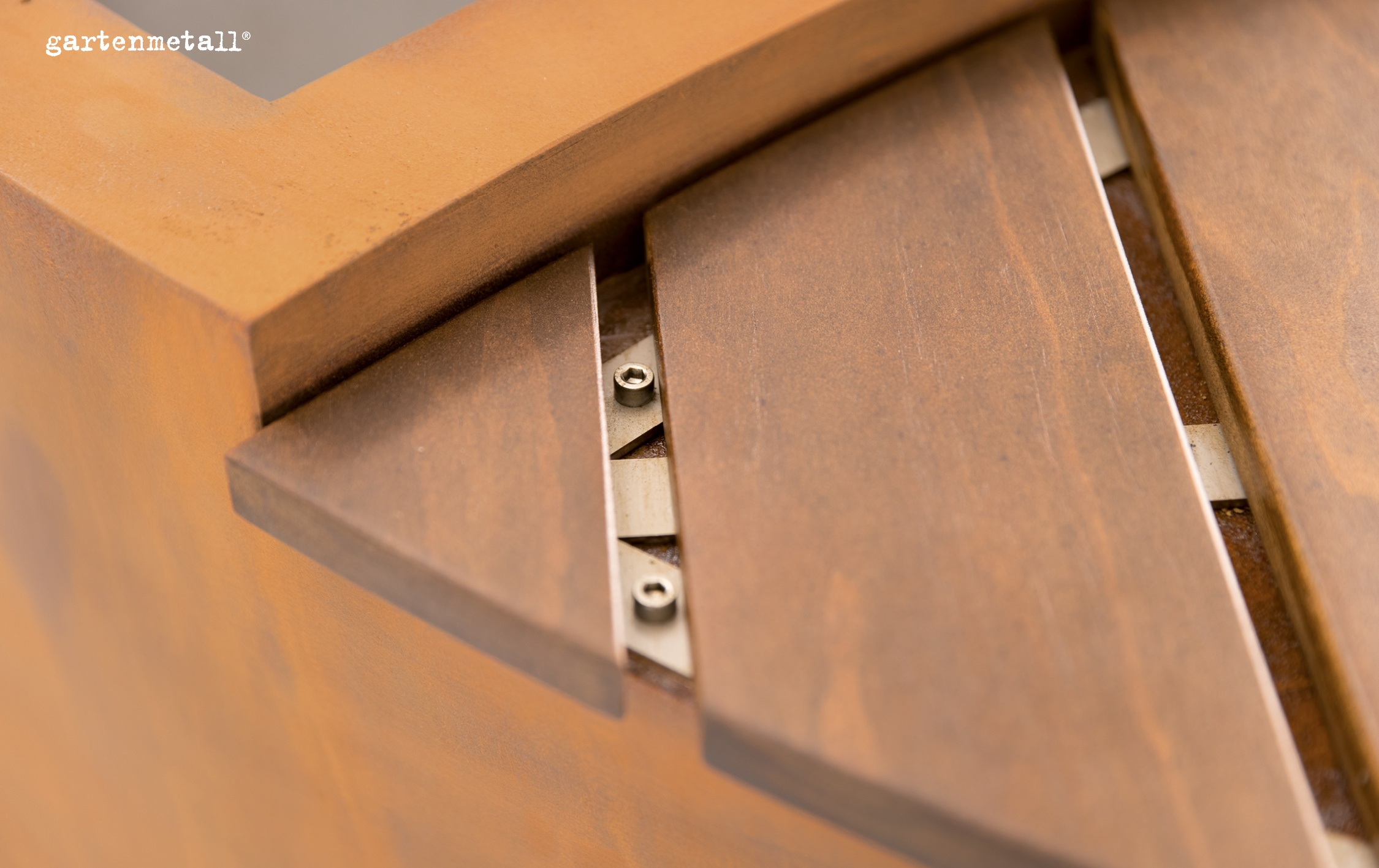 Metallverkleidung mit sichtbaren Schrauben und strukturiertem, rostbraunem Finish. Im Hintergrund ist ein Winkel zu erkennen. Oben links steht 'gartenmetall®'.