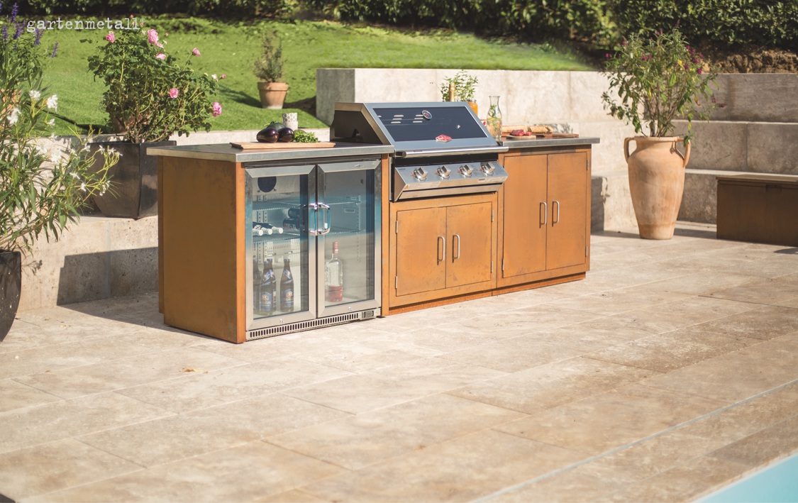 Außenküche mit Grill und Kühlschrank steht auf einer Terrasse, umgeben von Pflanzen und Gras. Oben links steht 'gartenmetall'. Sie ist aus rostbraunem Material gefertigt.