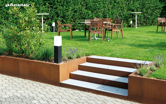 Gartenmöbel stehen auf einem gepflegten Rasen, im Vordergrund führen Treppenstufen zwischen Hochbeeten aus rostigem Metall hinunter auf einen gepflasterten Weg, Hecken bilden den Hintergrund. Text: 'gartenmetall®'.