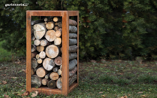 Holzscheite stapeln in einem rostfarbenen Metallrahmen, stehend auf grasbewachsenem Boden vor einem Hintergrund aus dichten, grünen Sträuchern. Oben links steht 'gartenmetall®'.