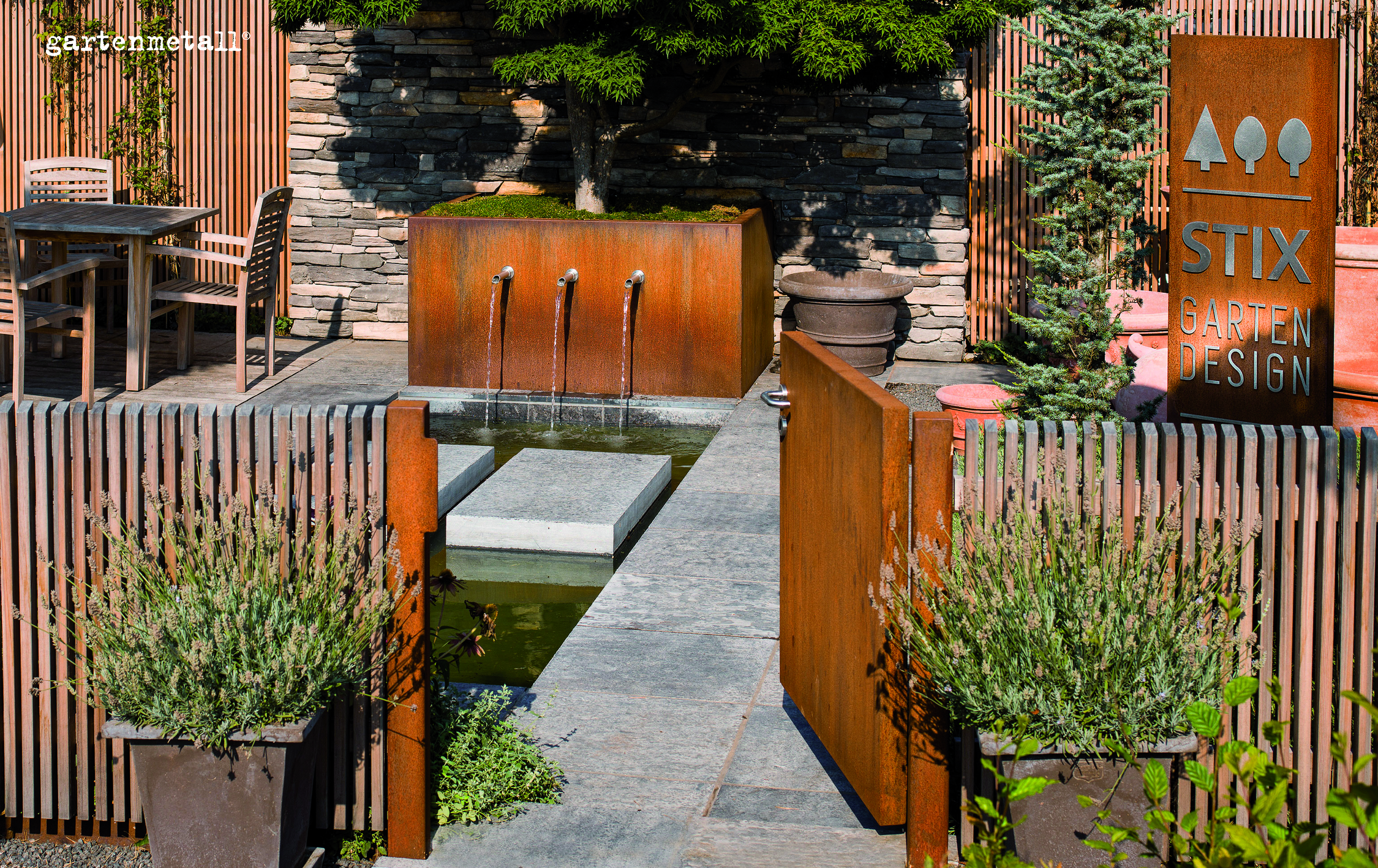 Gargoyles flow into a rectangular pond, surrounded by wooden fences and stone walls. Table with chairs on the left. Sign with 'Stix Garden Design' and 'gartenmetall' at the top left of the picture.