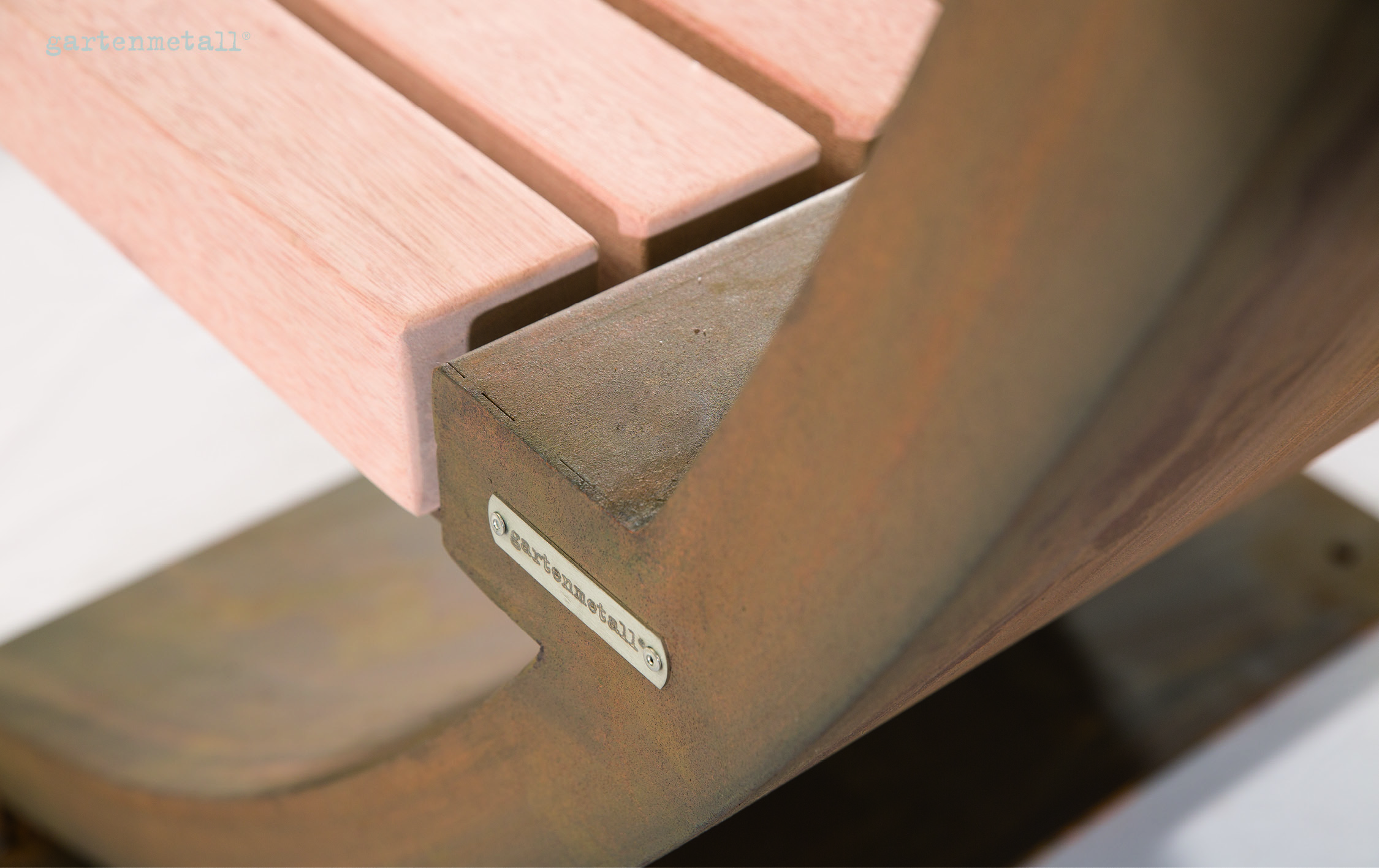 Holzbankdetail zeigt Metallrahmen mit graviertem Schild „gartenmetall“. Ruhend auf glattem Boden in heller Umgebung.