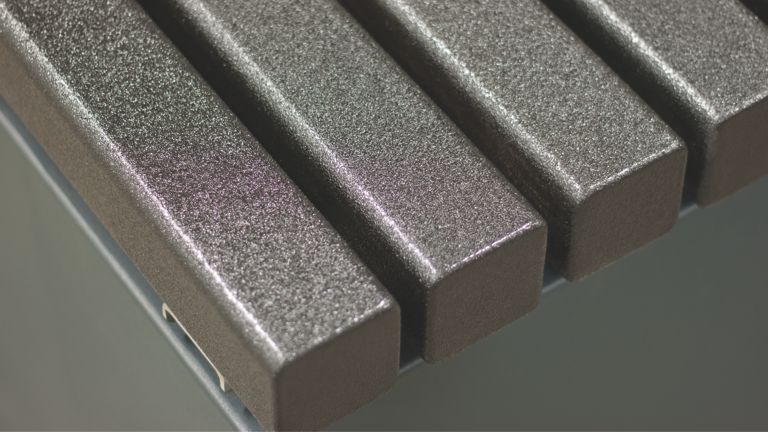 Metallstreben liegen parallel nebeneinander, bilden Sitzfläche, während sie auf einer Metallkonstruktion montiert sind. Die Oberfläche ist strukturiert und glitzert leicht im Licht.