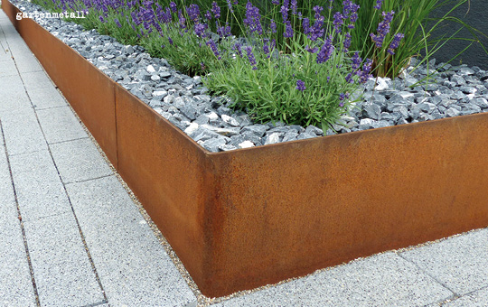 Rostfarbenes Metallbeet umrahmt Lavendel, umgeben von grauem Schotter, auf einer Pflastersteinfläche. In der oberen linken Ecke steht der Text 'gartenmetall'.