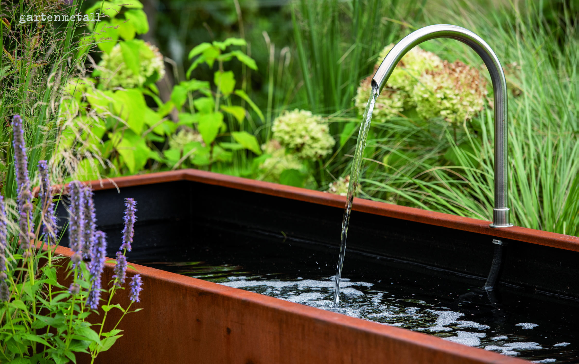Ein Stahlrohr fließt Wasser in ein rechteckiges Becken, umgeben von grünen Pflanzen. Text: 'gartenmetall' oben links.