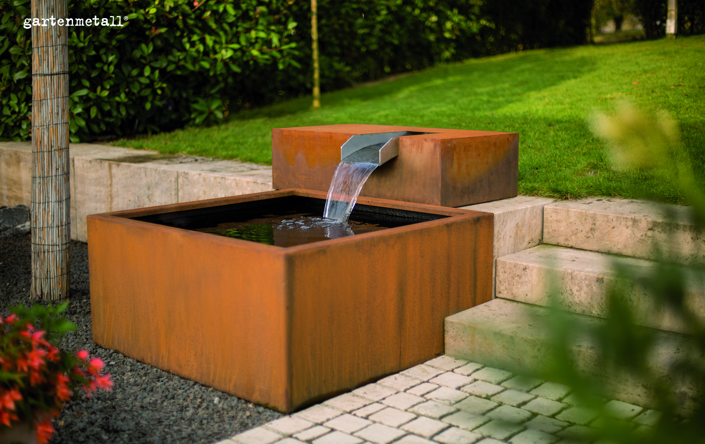 Wasser strömt aus einem Metallbrunnen in ein Becken. Der Brunnen steht auf einer Terrasse mit Steinfliesen und umgeben von gepflegtem Rasen. Text: 'gartenmetall®'.