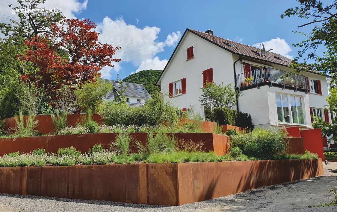 Ein terrassierter Garten mit Pflanzen auf rostfarbenen Stufen vor einem zweistöckigen Haus mit weißen Wänden und roten Fensterläden bei sonnigem Wetter. Text: „garteneinkauf“.