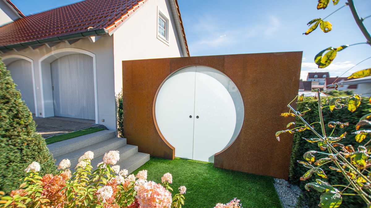 Runde Eingangstür geöffnet, umgeben von rostbrauner Mauer, führt in modernen Garten mit grünem Rasen. Haus mit rotem Ziegeldach und Blumen im Vordergrund.