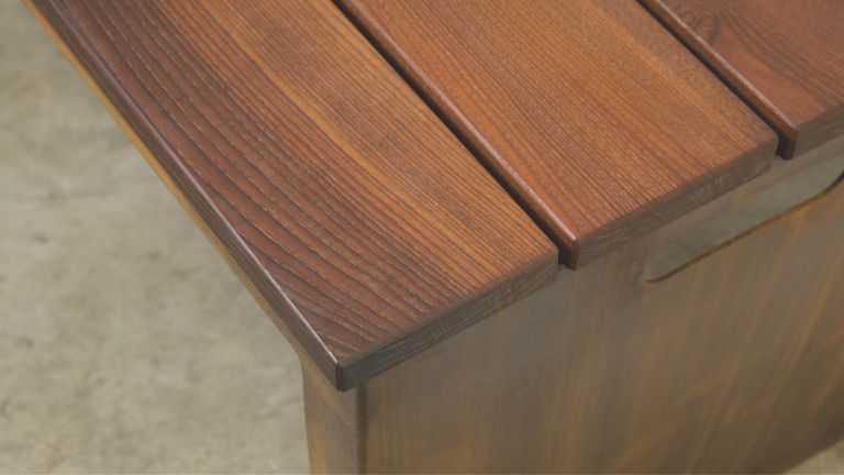 Ein brauner Holztisch mit klar sichtbarer Maserung steht still auf einem hellgrauen, strukturierten Untergrund, das Holz wirkt frisch bearbeitet und poliert. Kein Text vorhanden.