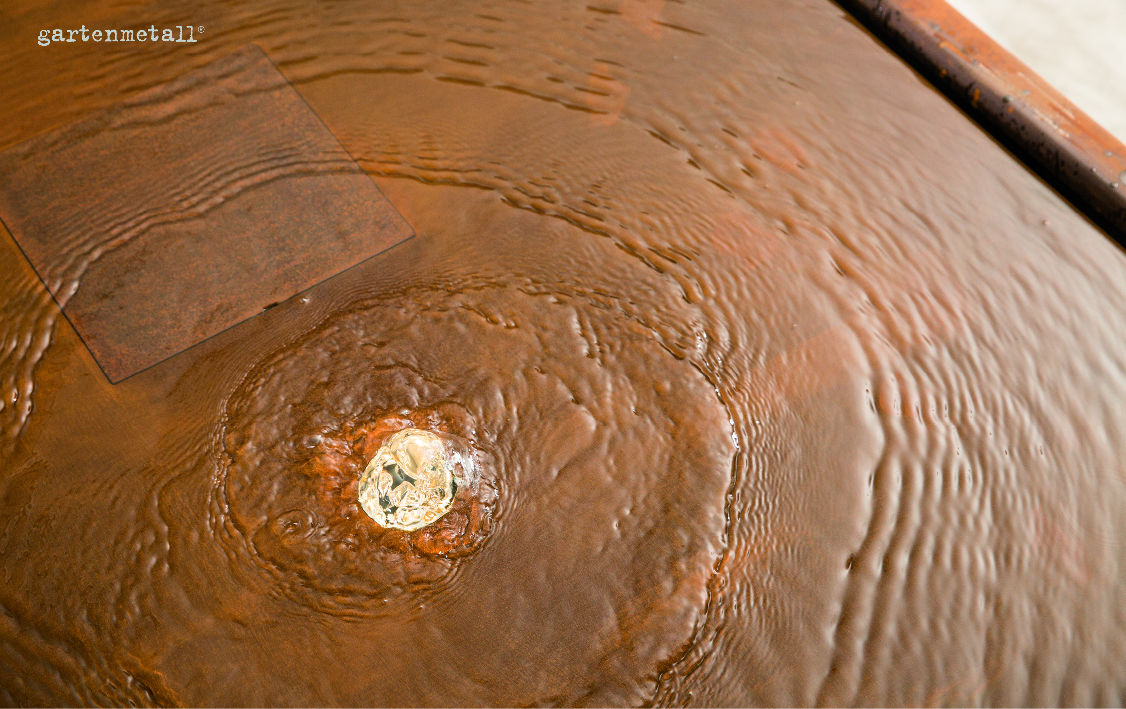 Wassertropfen spritzt aus einer metallischen Oberfläche, erzeugt konzentrische Wellen. Oberseite mit Rostoptik. Aufschrift oben links: 'gartenmetall®'. Umgebung unsichtbar, Fokus auf Wasserbewegung.